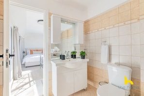 A modern bathroom with a white sink, mirror, and toilet. There is a bed visible through an open door in the adjacent room. - 3-Storey Family Home, City View & Private Parking! (Athina)