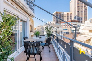 A balcony with a table and chairs, overlooking a cityscape with modern buildings. - 3-Storey Family Home, City View & Private Parking! (Athina)