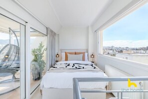 A modern bedroom with a large bed, a view of the city, and a balcony with a chair. - 3-Storey Family Home, City View & Private Parking! (Athina)