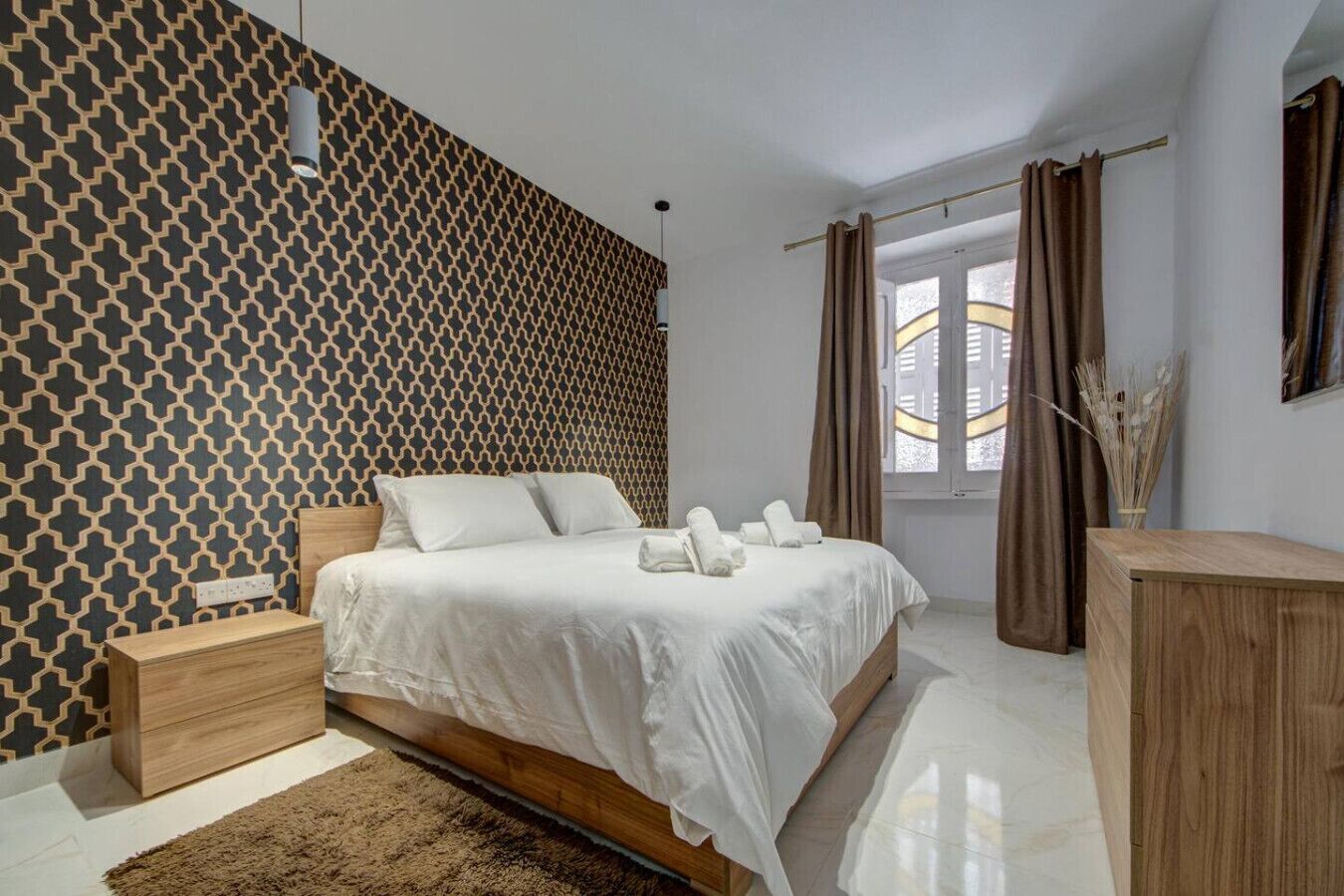 A modern bedroom with a geometric patterned wall, a wooden bed frame, a bedside table, and a nightstand.