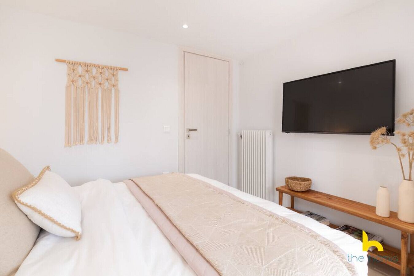 Un dormitorio con cama, televisor montado en la pared, radiador y mesita de noche con cesta y velas.