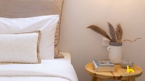 A neatly made bed with white linens and a decorative pillow, a small wooden side table with a book and a vase of dried plants, and a wall-mounted light fixture. - Rooftop Hideaway w/ Large Private Patio near Metro (Athina)