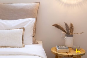 A neatly made bed with white linens and a decorative pillow, a small wooden side table with a book and a vase of dried plants, and a wall-mounted light fixture. - Rooftop Hideaway w/ Large Private Patio near Metro (Athina)