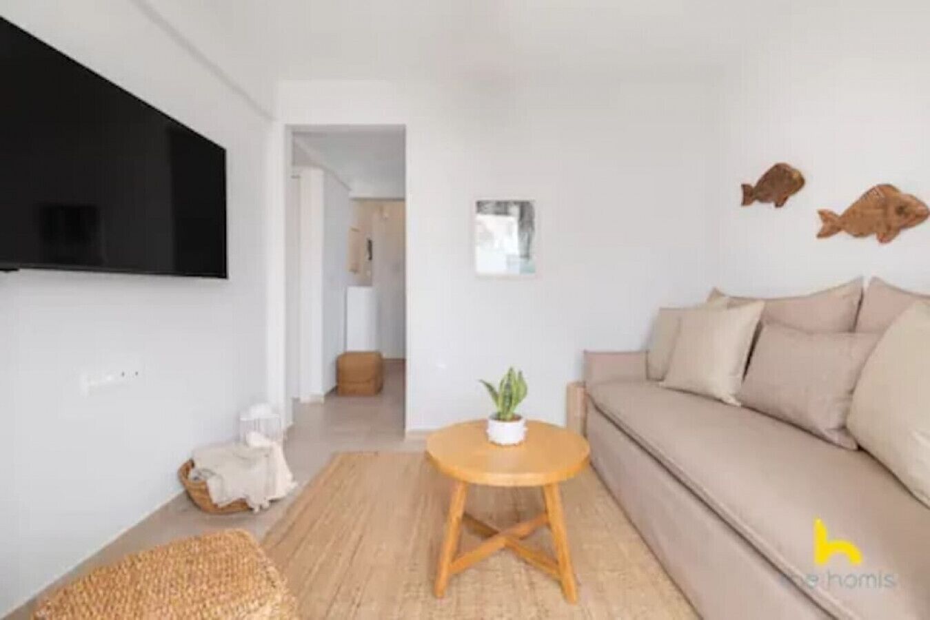 Un salon avec un canapé beige, une petite table ronde en bois sur laquelle est posée une plante, et un téléviseur fixé au mur.