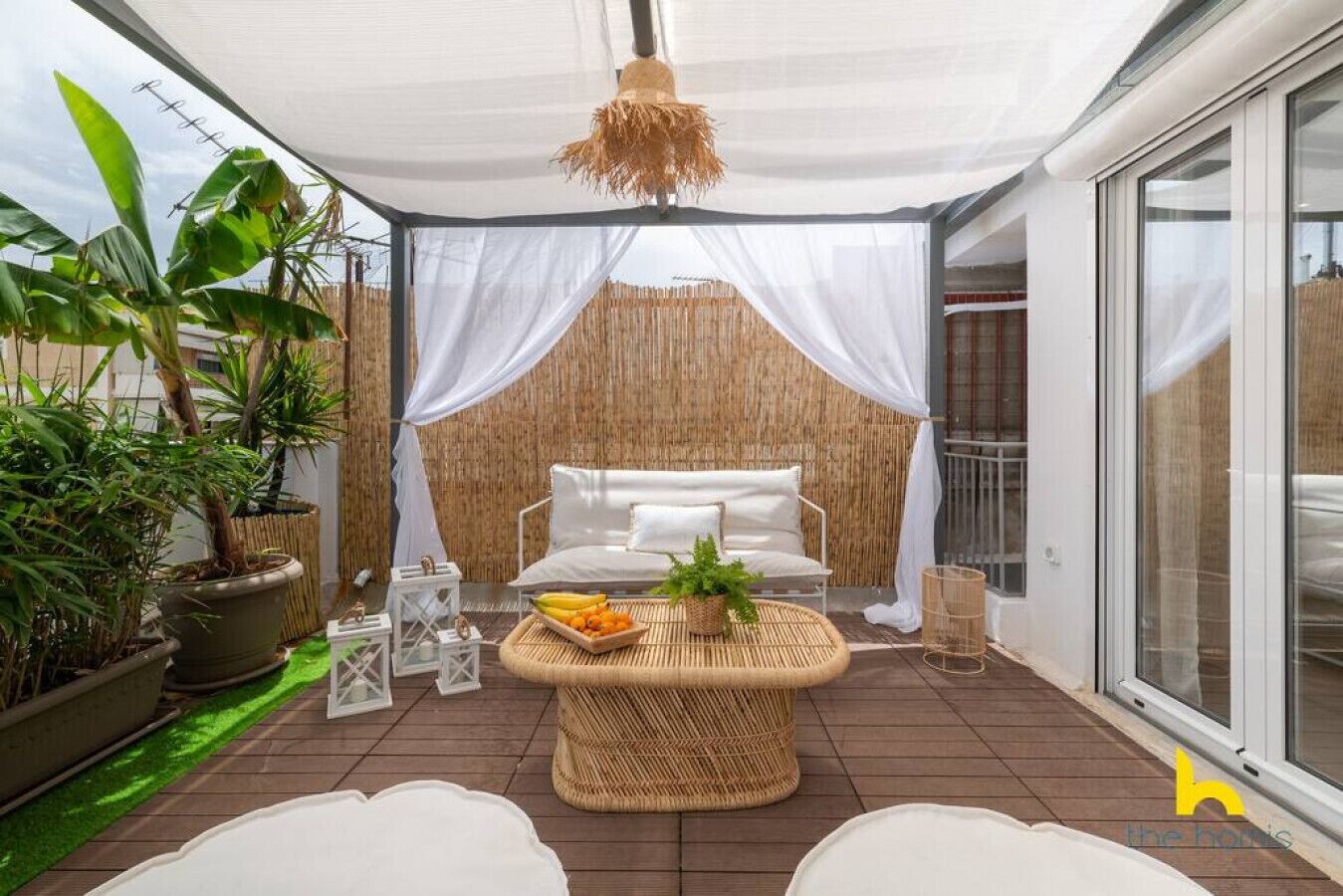 A covered patio area with a white sofa, a wicker coffee table with fruits and a plant, potted plants, and a bamboo wall.