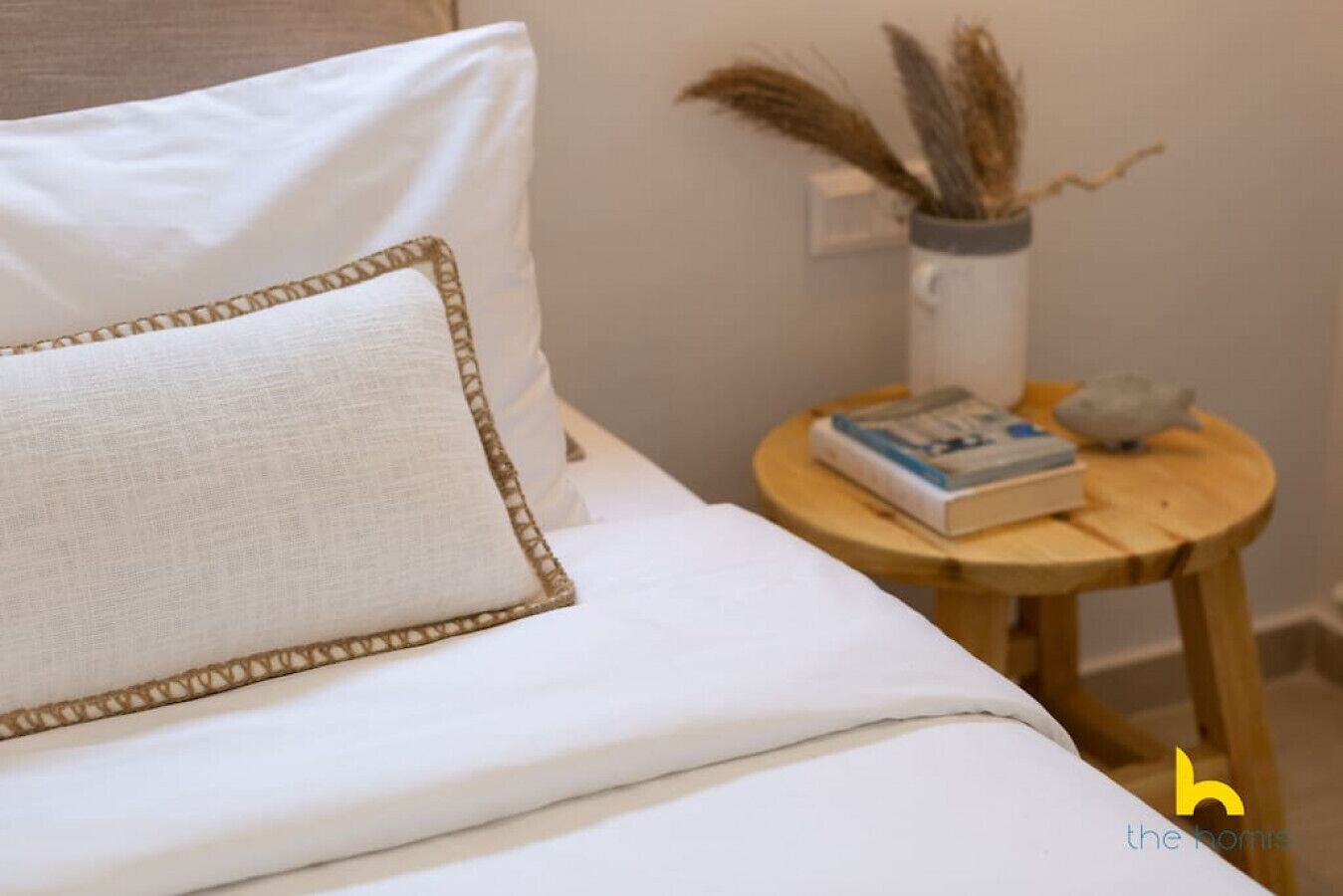 Una cama bien hecha con un almohadón blanco y un cojín decorativo con ribete dorado. Junto a la cama, una mesita de madera con un libro y un jarrón con plantas secas.