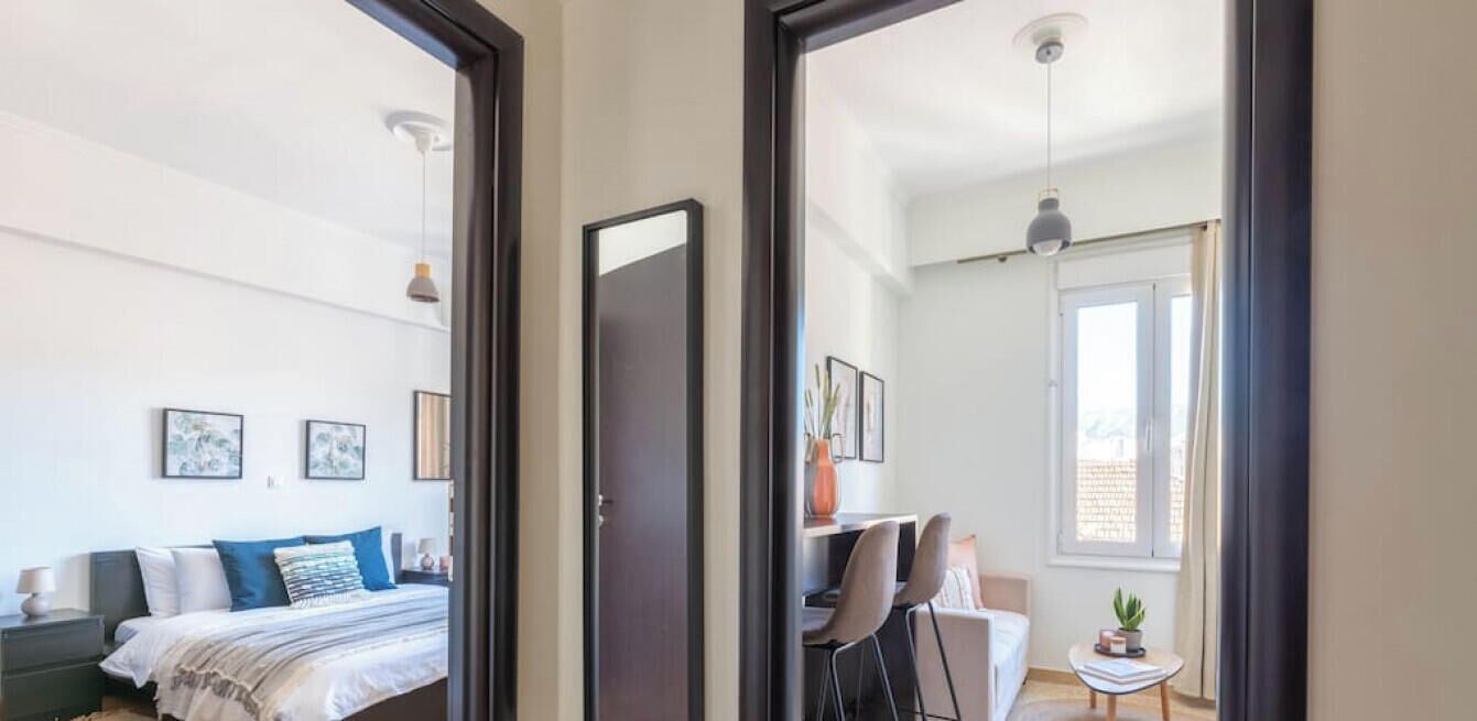 Quarto com cama, mesa de cabeceira e escrivaninha com cadeira. Há uma janela com cortinas e vista para o exterior.
