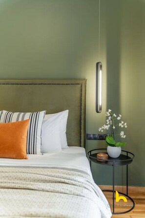 A neatly made bed with striped pillows and a single orange pillow, a bedside table with a potted orchid, and a hanging light fixture. - Top-Floor Oasis, King-Bed & Whirlpool, Acropolis! (Athina)
