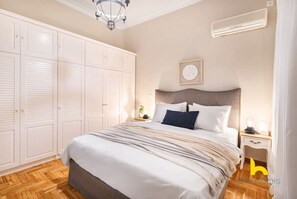 A bedroom with a large bed, a wardrobe, a bedside table with a lamp, and a wall clock. - Grand Family APT with Courtyard near Acropolis! (Athina)