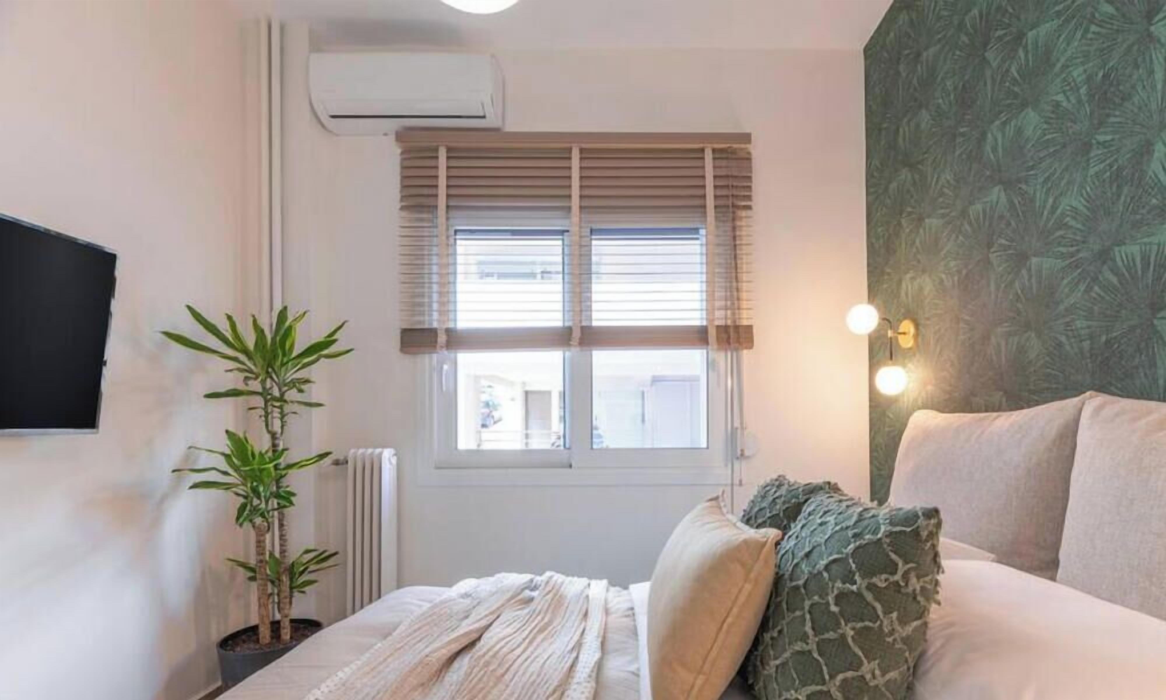A bedroom with a bed, a potted plant, a radiator, a window with blinds, and a wall-mounted TV.