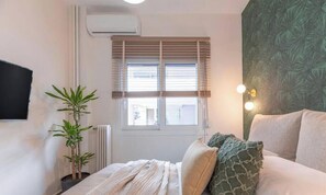 A bedroom with a bed, a potted plant, a radiator, a window with blinds, and a wall-mounted TV. - The Urban Jungle Escape Corner APT Near Acropolis (Athina)