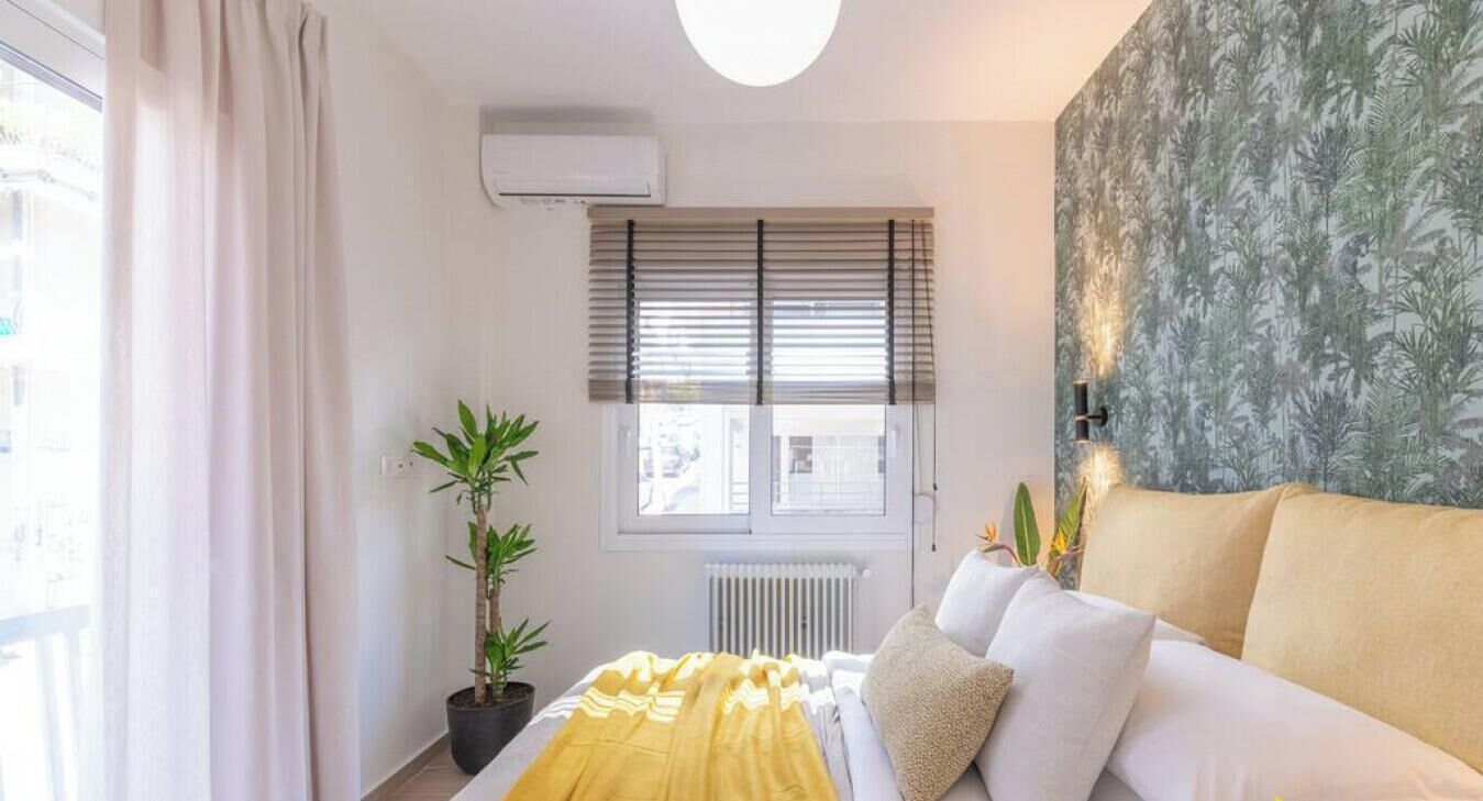 Une chambre à coucher avec un lit, une fenêtre avec des stores, un radiateur, une plante en pot et un mur orné d’un motif de feuilles.