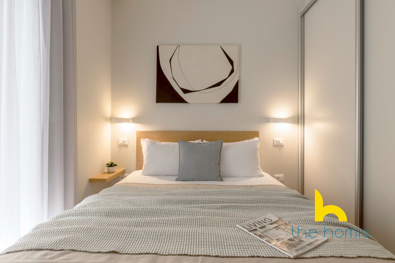 Une chambre d’hôtel bien rangée, avec un lit, une table de chevet dotée d’une lampe, une œuvre d’art accrochée au mur et un magazine posé sur le lit.