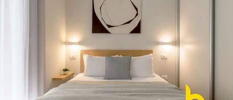A neatly arranged hotel room with a bed, a bedside table with a lamp, a wall-mounted artwork, and a magazine on the bed.
