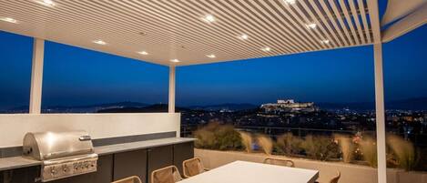 Terrassa exterior amb una barbacoa, taula de menjador i cadires, amb vista a un paisatge urbà i un turó amb una estructura semblant a un castell.