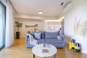 A modern living room with a grey sofa, a white coffee table, a potted plant, and a kitchen area in the background. - Urban APT with Stunning Acropolis View & Roofdeck (Athina)