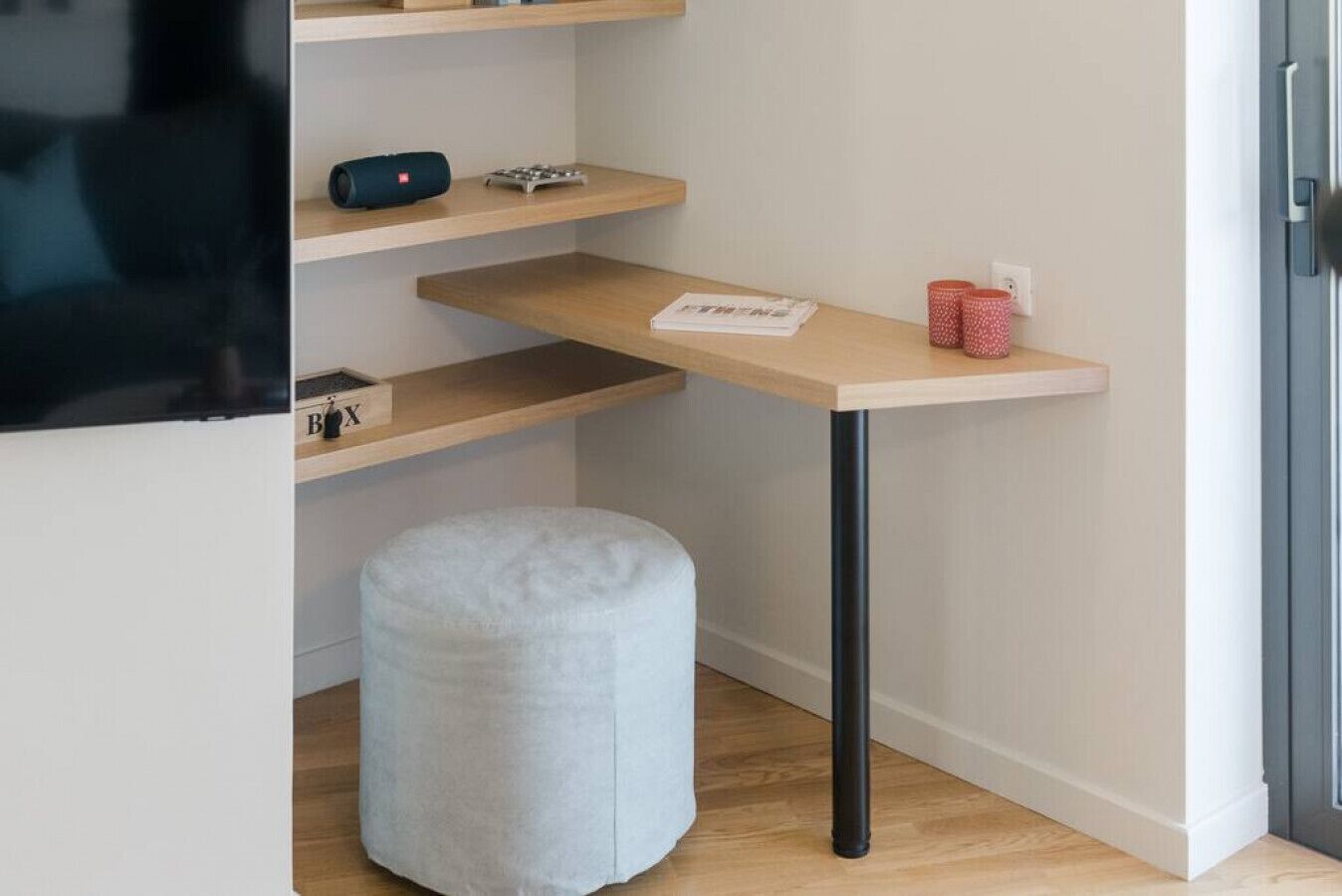 Een kleine, moderne kamer met een houten bureau, een witte poef en een aan de muur bevestigde televisie.