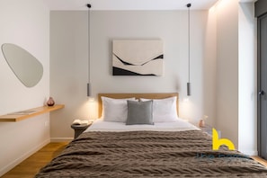 Une chambre moderne avec une tête de lit en bois, un grand lit recouvert d’une couverture à motifs, des tables de chevet avec des lampes, un tableau abstrait accroché au mur et un miroir sur le mur de gauche.