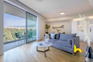 A modern living room with a grey sofa, a coffee table, and a view of a cityscape through large windows. - Urban APT with Stunning Acropolis View & Roofdeck (Athina)
