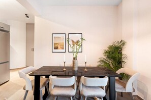 Ein modernes Esszimmer mit einem dunklen Holztisch, weißen Polsterstühlen, zwei gerahmten Bildern an der Wand und einem Blumentopf.