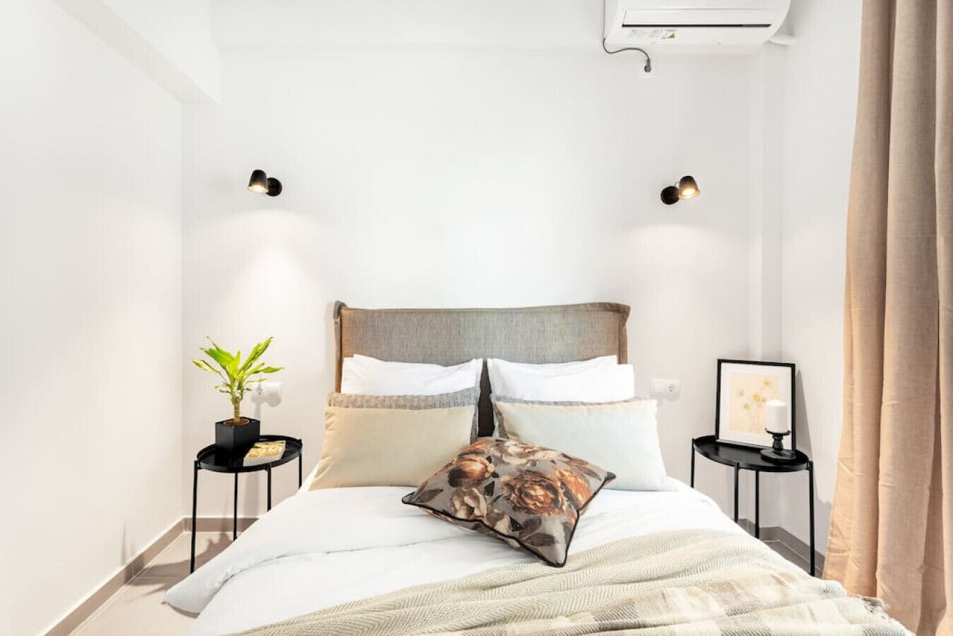 A neatly arranged bedroom with a bed, two bedside tables, a potted plant, and a wall-mounted air conditioner.