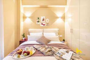 A neatly made bed with pillows, a bedside table with a plant, and wall-mounted artwork. - Quiet, Minimalist Base w/ Balcony near Metro (Athina)