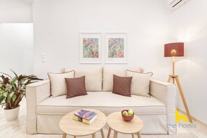 A modern living room with a beige sofa, two patterned throw pillows, a small round table with books and a bowl of fruit, a potted plant, and a floor lamp. - Quiet, Minimalist Base w/ Balcony near Metro (Athina)