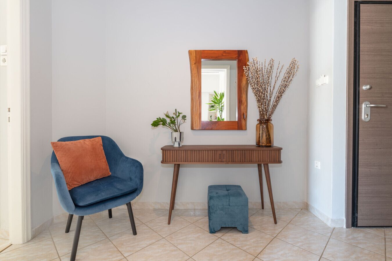Un interior moderno con un sillón azul, un taburete pequeño, una consola de madera, un espejo y un jarrón con plantas secas.