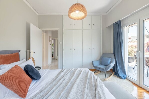 Un dormitorio con cama, una silla, un armario y vistas a través de un ventanal.