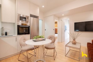 Cozinha moderna com eletrodomésticos de aço inoxidável, área de refeições com mesa redonda e quatro cadeiras, e uma pequena mesa lateral com um vaso.