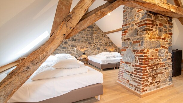 A bedroom with a sloped ceiling, exposed wooden beams, and a stone wall. There are two beds with white bedding. - Charming holiday home with garden (Dinant)