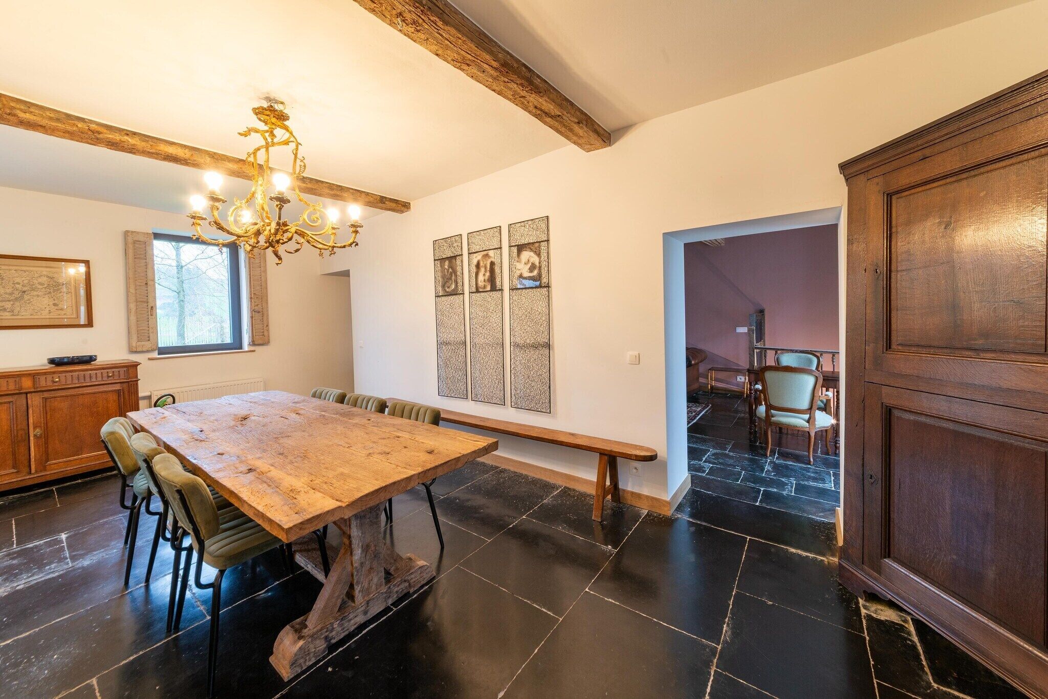 A dining area with a wooden table and chairs, a bench, a chandelier, and a view of a room with a chair and a cabinet.