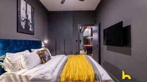 A modern bedroom with a tufted blue headboard, a bed with white linens and a yellow throw, a ceiling fan, a framed picture on the wall, and a TV mounted on the wall. - Unique Urban Cave w/ Pool Table Near Acropolis! (Athina)