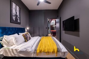 A modern bedroom with a tufted blue headboard, a bed with white linens and a yellow throw, a ceiling fan, a framed picture on the wall, and a TV mounted on the wall. - Unique Urban Cave w/ Pool Table Near Acropolis! (Athina)