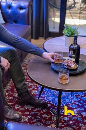 A person seated at a round table with a bottle and glasses, a patterned carpet, and a blue couch in the background. - Unique Urban Cave w/ Pool Table Near Acropolis! (Athina)