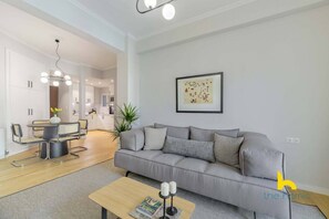 A modern living room with a grey sofa, a coffee table, and a dining area with a round table and chairs. - Sun-Splashed 2-BDR Corner APT with Hillside views! (Athina)