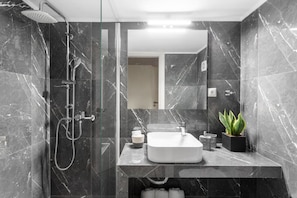 A modern bathroom with a glass-enclosed shower, a marble-tiled wall, a large mirror, and a potted plant.