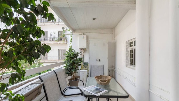A balcony with a table and chairs, a potted plant, and a view of the building across the street. - Comfortable & Quiet Family APT Near Acropolis (Athina)