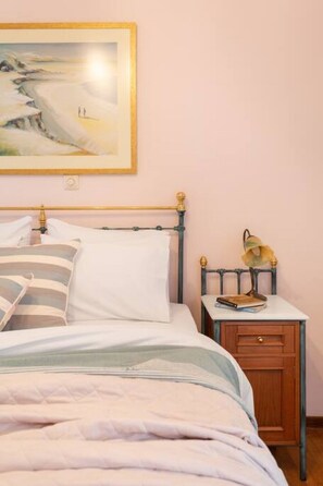 A neatly made bed with striped pillows, a bedside table with a lamp, and a painting on the wall. - Bright, Calming Retreat With Balcony Near Metro! (Menemeni)