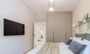 A bedroom with a bed, a TV mounted on the wall, a wardrobe, and a door leading to another room. - Minimal 2-BDR Apt Near Acropolis By The Homis (Athina)