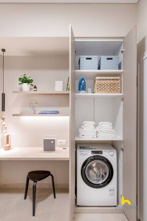 A laundry room with a washing machine, shelves with folded towels, a potted plant, and a desk with a chair. - Minimal 2-BDR Apt Near Acropolis By The Homis (Athina)