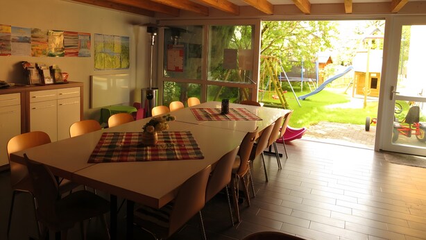 A dining area with a table, chairs, and a view of a playground through large windows. - Apartment '2' with Balcony, Garden and Wi-Fi (Überlingen)