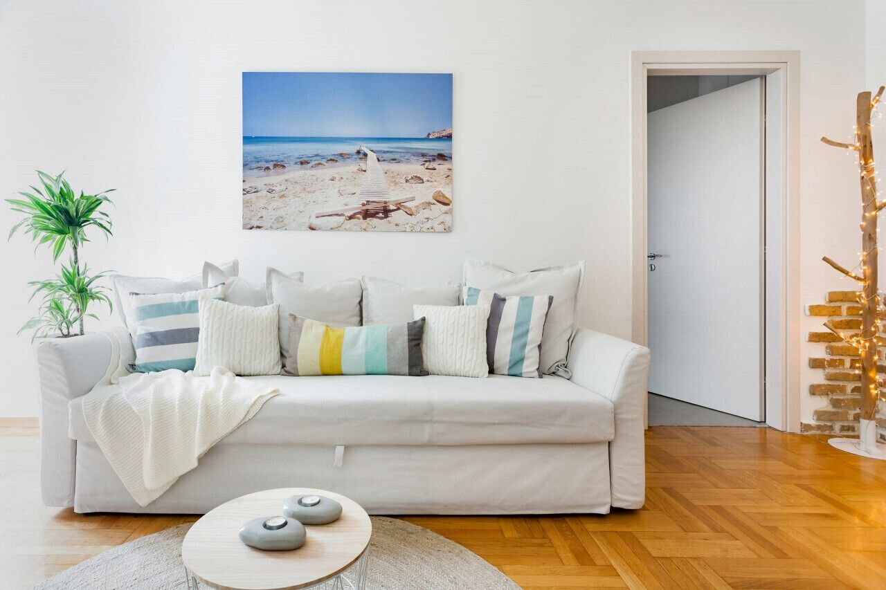 A living room with a white sofa, a painting of a beach, a potted plant, and a decorative tree.