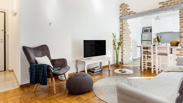A modern living room with a flat-screen TV, a comfortable armchair, a round ottoman, and a dining area with a white table and chairs. - Head for Acropolis from a Romantic Nest in Koukaki (Athina)