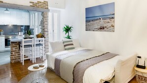 A compact room with a kitchenette, a bed, a dining area, and a beach-themed wall art. - Head for Acropolis from a Romantic Nest in Koukaki (Athina)