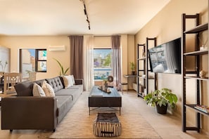 Uma sala de estar moderna com sofá, mesa de centro e televisão. Há uma varanda com vista para edifícios.