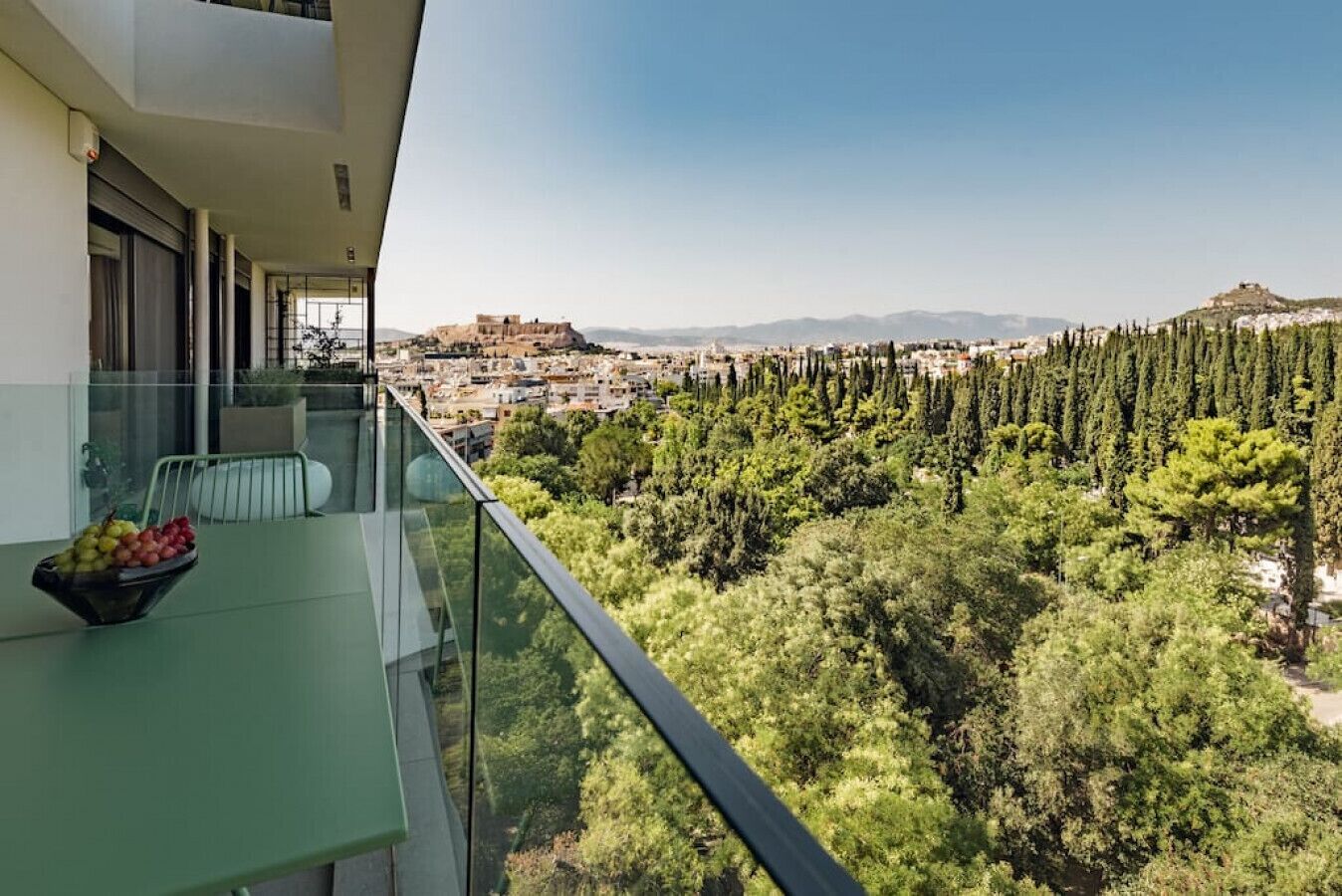 A balcony with a glass table and a bowl of fruit, overlooking a cityscape with trees and a distant hill.