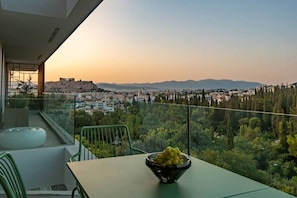 A balcony with a table and chairs, a bowl of fruit, and a view of a cityscape and mountains. - Kalypso, Urban APT w/ Unique Views of Acropolis! (Athina)
