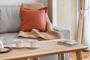 A cozy living room with a grey sofa, a red patterned pillow, a beige blanket, and a wooden coffee table with cups and a book. - Chill Out In Retropolitan Hideaway at Alsos Park (Athina)