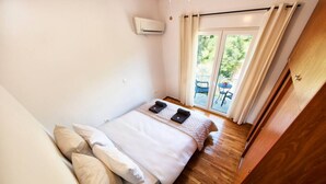 A bedroom with a bed, pillows, a bedside table, an air conditioning unit, and a view of the outdoors through a glass door. - Acropolis Hillside Escape, Sea & Nature Views (Athina)